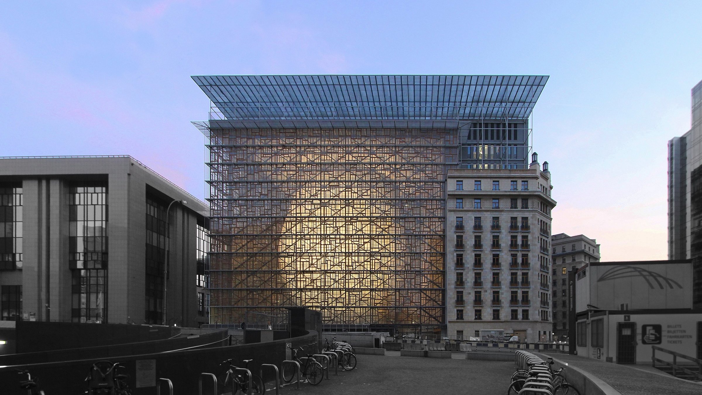 L'image montre un bâtiment moderne avec une structure en verre et en métal, se dressant au centre d'un environnement urbain. La façade réfléchissante crée un effet de transparence et de lumière, tandis que le ciel présente des nuances de bleu et de rose, suggérant un moment au crépuscule. Au premier plan, on aperçoit des vélos alignés, ajoutant une touche de vie à la scène. À gauche, il y a un autre bâtiment avec un style architectural plus traditionnel, contrastant avec la modernité du bâtiment principal.