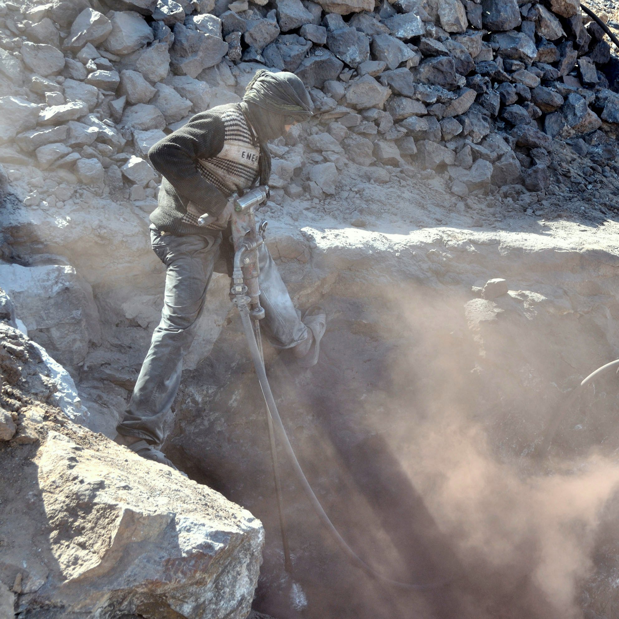 L'image montre un mineur en train d'extraire des matériaux dans une carrière. Il est vêtu de vêtements de protection et cache son visage avec un foulard, probablement pour se protéger de la poussière. La scène est poussiéreuse, et des débris de roches sont visibles autour de lui. Le mineur utilise un outil, probablement un marteau-piqueur, pour travailler dans la roche. L'environnement est aride et rocheux, typique des sites miniers.