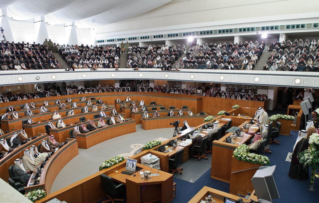 L'image montre une grande assemblée, probablement un parlement ou une salle de réunion officielle. On y voit des hommes en tenue traditionnelle, souvent vêtus de thobes et de keffiehs, assis à des bureaux en bois agencés en demi-cercle. La salle est bien aménagée, avec des décorations florales sur les tables et des écrans affichant des informations. Une grande audience est présente, ce qui suggère une session importante. L'ambiance semble solennelle et axée sur la discussion ou la prise de décision.