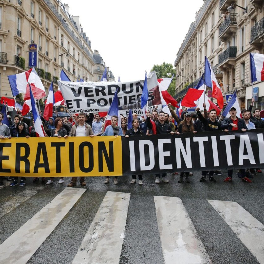 L'image montre une manifestation dans une rue, où un groupe de personnes marche en brandissant des drapeaux français. Ils tiennent une grande banderole avec les mots "GENERATION IDENTITAIRE". En arrière-plan, d'autres banderoles sont visibles, affichant des slogans supplémentaires. L'atmosphère semble énergique et engagée, avec des participants portant des vêtements variés. Les bâtiments en arrière-plan reflètent un cadre urbain typique.