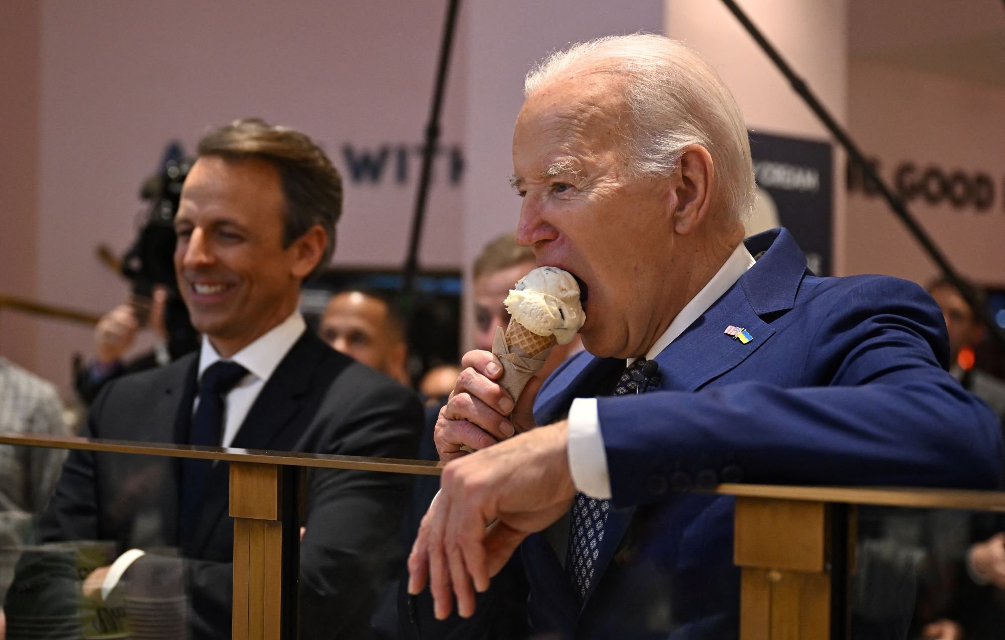 الصورة تُظهر رجلًا في بدلة رسمية وهو يأخذ قضمة من مثلجات، بينما يقف بجانبه رجل آخر يبتسم. يبدو أن المكان عبارة عن حدث أو اجتماع، حيث يوجد العديد من الأشخاص في الخلفية. الرجل الذي يأكل المثلجات هو شخص بارز يبدو سعيدًا وهو يستمتع باللحظة.