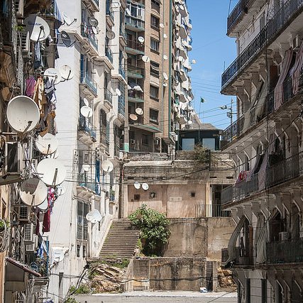 L'image montre une vue urbaine d'une rue avec des bâtiments en pierre et des murs en béton. On observe un mélange d'architecture ancienne et moderne, avec des balcons ornés de fer forgé. De nombreux paraboles satellites sont installées sur les façades des bâtiments, tandis que des linges pendus à l'extérieur ajoutent une touche de vie quotidienne. Les escaliers menant à d'autres niveaux sont visibles, et le ciel est clair et ensoleillé, donnant une atmosphère vive à la scène.