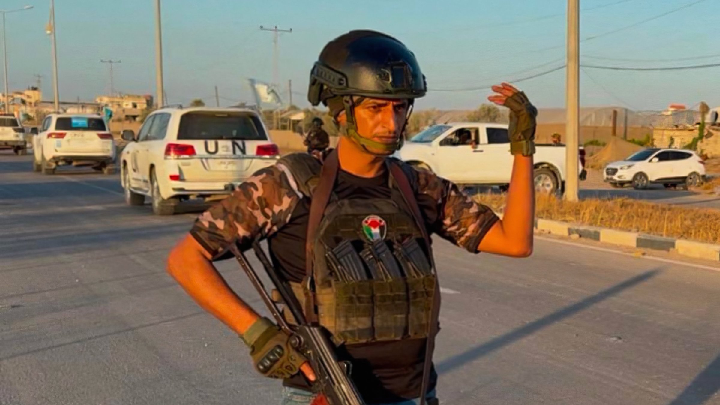 یک مرد با کلاه ایمنی و جلیقه ضد گلوله، اسلحه کلاشنیکف را در دست دارد و در خیابان ایستاده است.