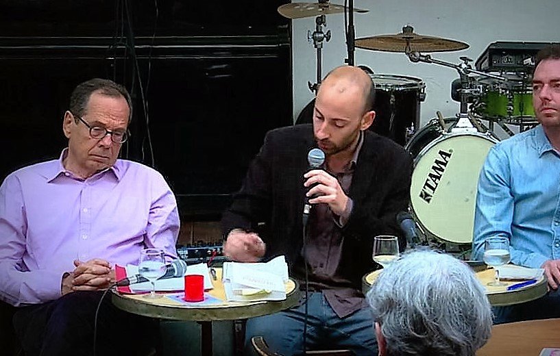 L'image montre un moment lors d'un événement où trois hommes sont assis autour de petites tables. Au centre, un homme parle dans un micro, semblant engager une discussion ou une présentation. Les autres hommes, à sa gauche et à sa droite, écoutent attentivement. On peut voir des instruments de musique en arrière-plan, ce qui suggère que l'événement se déroule dans un cadre décontracté, peut-être un café ou un bar. Des verres et des notes sont présents sur les tables, ajoutant à l'ambiance informelle de la rencontre.
