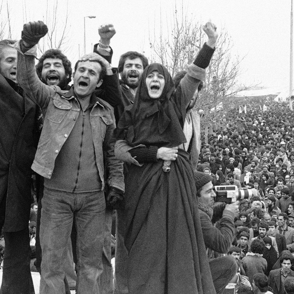 L'image montre un rassemblement de personnes, probablement lors d'une manifestation historique. Au premier plan, un groupe de manifestants lève les poings en l'air, exprimant leur enthousiasme ou leur détermination. Certains membres du groupe portent des vêtements traditionnels, tandis que d'autres sont habillés de manière plus moderne. À l'arrière-plan, une grande foule est visible, rassemblée autour d'un monument emblématique, ce qui suggère l'importance de l'événement. L'atmosphère semble chargée d'énergie et de passion, reflétant un moment significatif de mobilisation collective.