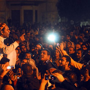 L'image montre une grande foule rassemblée dans une ambiance nocturne. Un homme au centre, visiblement passionné, est en train de parler ou de s'exprimer avec intensité. Les visages de la foule sont éclairés par des lumières, peut-être des téléphones portables ou des caméras. Les expressions des gens varient entre l'engagement, la curiosité et l'émotion, suggérant un moment de rassemblement ou de protestation. L'ensemble dégage une atmosphère vibrante et dynamique.