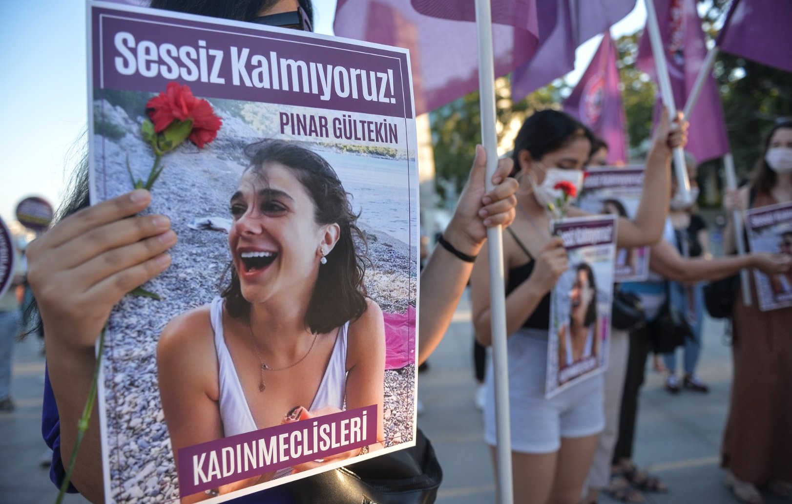 L'image montre un groupe de personnes manifestant avec des pancartes. L'une des pancartes porte le visage d'une jeune femme, accompagnée d'un message en turc qui se traduit par "Nous ne restons pas silencieuses !". Une manifestante tient une fleur rouge. Les femmes portent des vêtements variés et semblent engagées dans une cause, probablement liée à la défense des droits des femmes. L'atmosphère peut être perçue comme déterminée et solidaire, avec un accent sur la mémoire d'une personne nommée Pınar Gültekin.