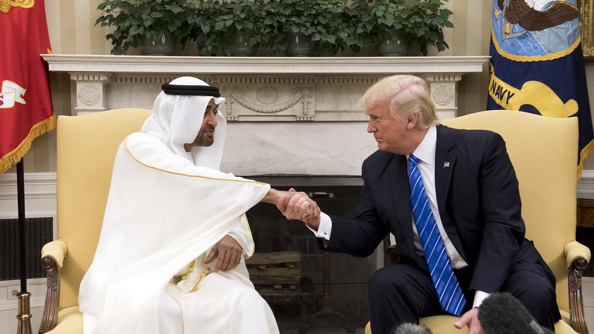L'image montre deux hommes en train de se serrer la main dans un bureau. L'un porte une tenue traditionnelle, probablement un kandoura et un chapeau, tandis que l'autre est en costume avec une cravate bleue. Ils semblent être en discussion dans un cadre formel, probablement dans un bureau gouvernemental, avec des plantes et des drapeaux visibles en arrière-plan.