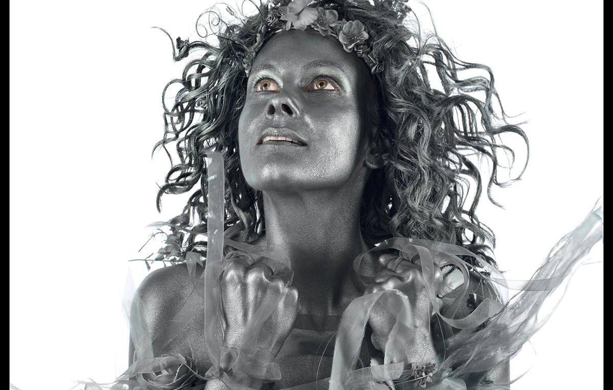 L'image affiche une femme peinte en argent, avec des cheveux ébouriffés et un regard intense. Elle est présentée dans un style dramatique, évoquant une performance artistique. Le fond est noir, ce qui met en avant la couleur métallique de son corps. En haut, on voit le titre de l'événement "D(rôles de printemps)" en grandes lettres, suivi des informations sur le lieu et les dates de l'événement, qui se déroule au TARMAC à Paris. Des détails supplémentaires sur les performances de théâtre et de danse sont également mentionnés, soulignant la diversité culturelle et artistique de l'événement.