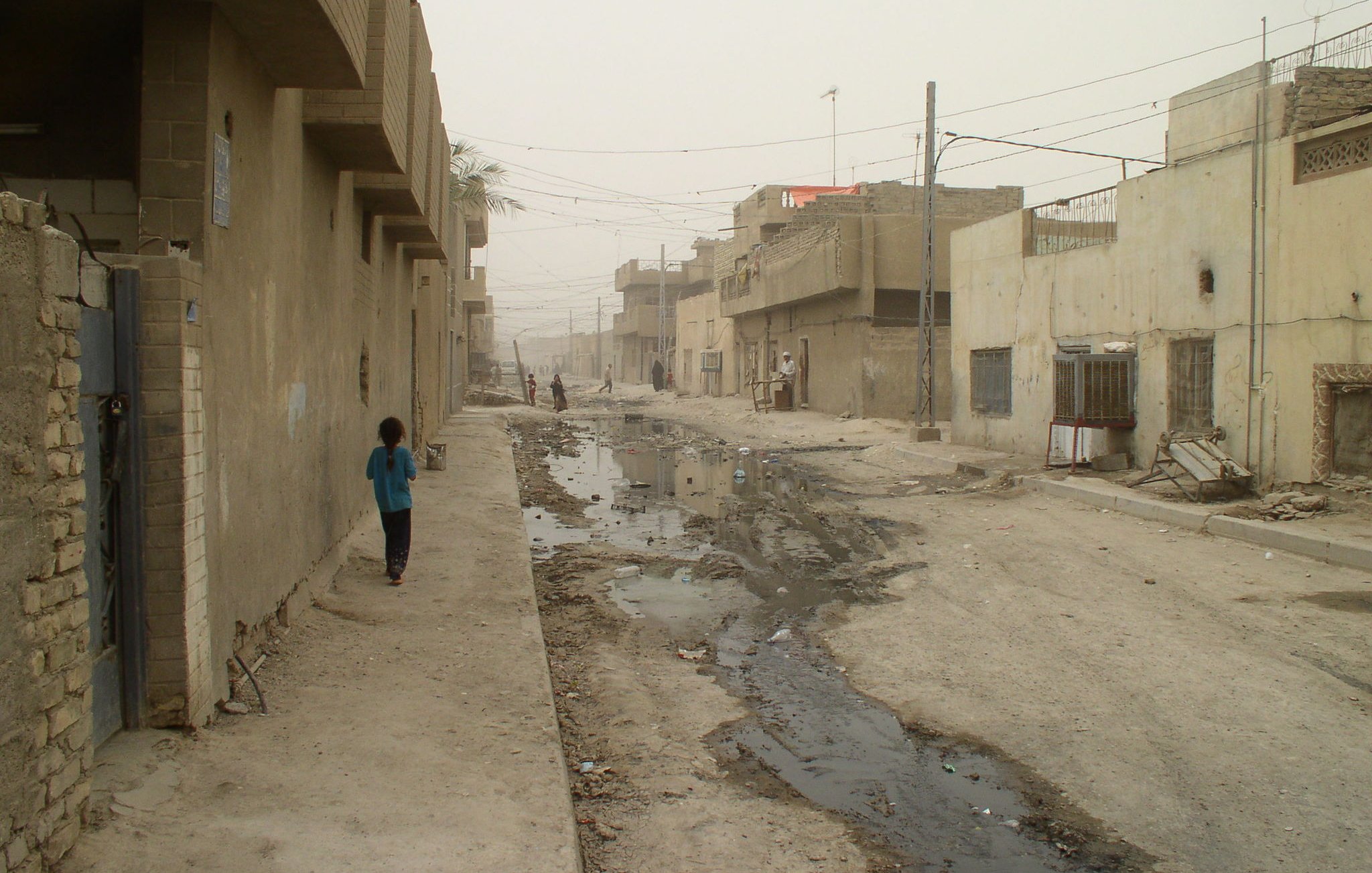 الصورة تظهر شارعًا في منطقة سكنية تبدو مهملة، حيث تظهر المباني ذات اللون الرمادي والخالي من الزينة. الطريق يبدو غير معبد ويظهر فيه أجزاء من المياه والأتربة. هناك شخصان يسيران في الشارع، ورغم الغبار، فإن تفاصيل الحياة اليومية تظهر في هذه البيئة. الشارع يبدو ضيقًا وفارغًا، مما يعكس حالة من الإهمال والبساطة في التصميم العمراني.