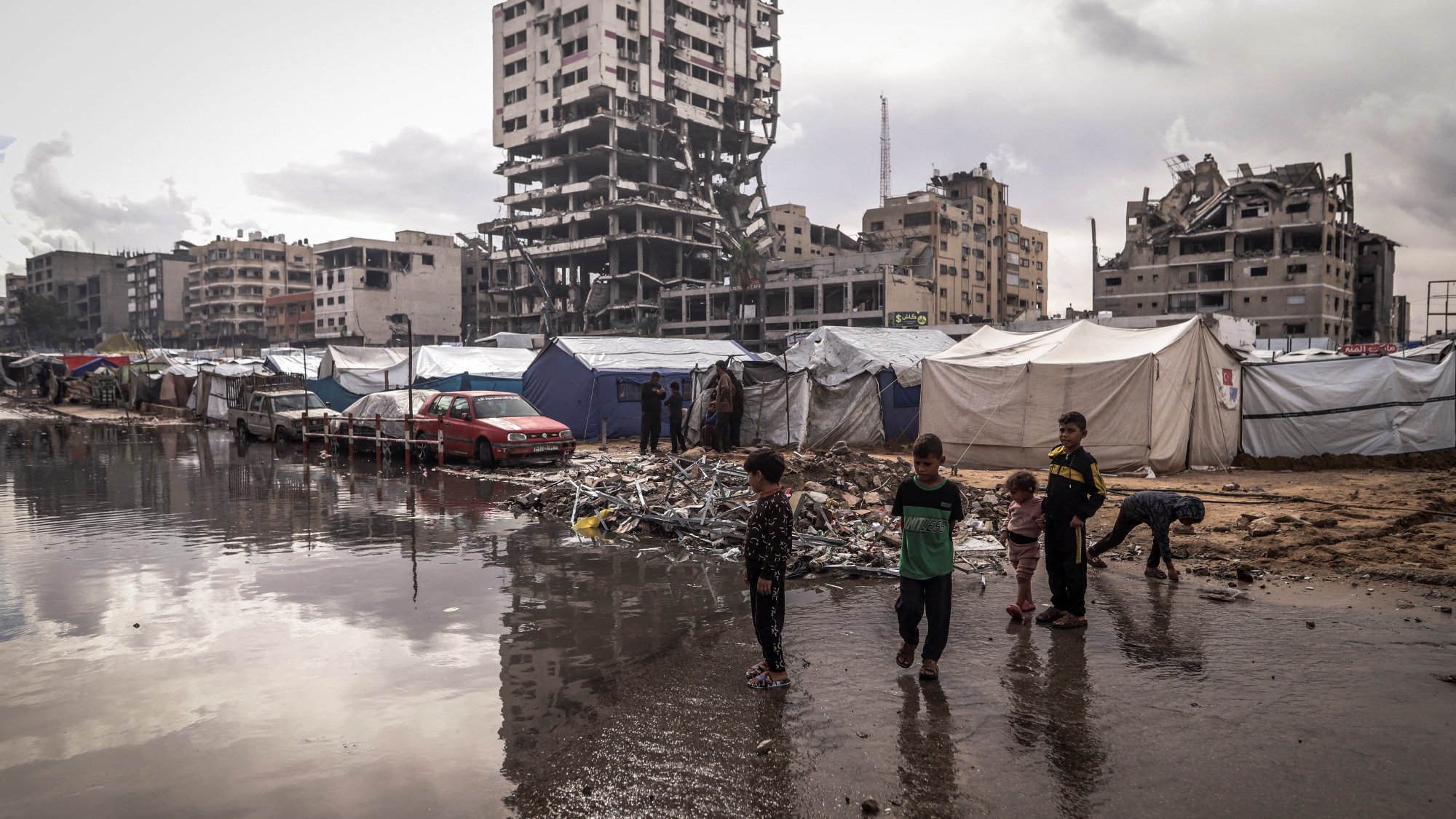 Un campo profughi con tende, bambini giocano e un edificio in rovina sullo sfondo.