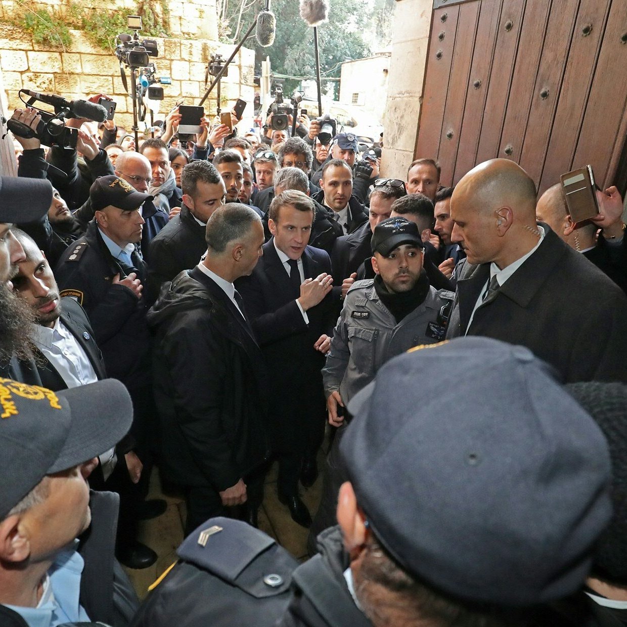 L'image montre une scène animée avec de nombreuses personnes rassemblées autour d'un groupe central. Des agents de sécurité, en uniforme, semblent encadrer des individus, probablement des personnalités importantes. L'atmosphère est tendue, avec des caméras et des journalistes présents, suggérant que cet événement pourrait avoir une importance médiatique. L'ensemble se déroule devant une grande porte, indiquant un lieu significatif, possiblement un bâtiment officiel ou historique.