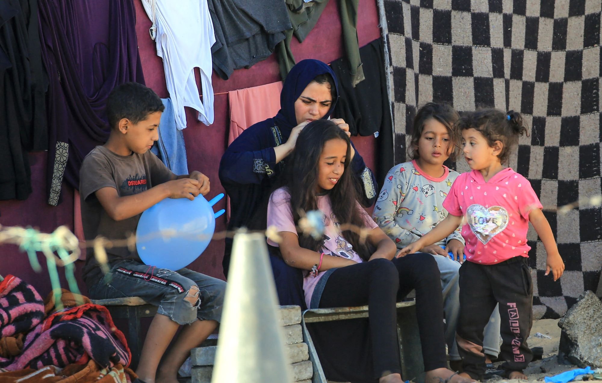 L'image montre un groupe d'enfants et une femme assis à l'extérieur, dans un environnement simple. Les enfants semblent interagir joyeusement, avec une petite fille qui parle à une autre. Une femme en vêtements traditionnels s'occupe des cheveux d'une jeune fille assise devant elle. En arrière-plan, on peut voir des vêtements accrochés et un sol en sable. L'ambiance est conviviale et émotive, évoquant des moments de partage et de complicité.