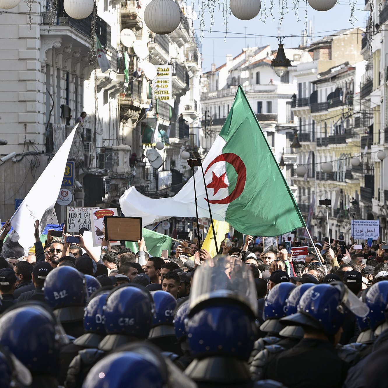 تظهر الصورة تجمعًا حاشدًا في شارع بالجزائر، حيث يحمل المتظاهرون الأعلام الجزائرية ويعبرون عن مطالبهم. يوجد في مقدمة الصورة رجال شرطة يرتدون خوذات واقية، وكأنهم يراقبون الوضع. يُلاحظ وجود لافتات ومحتجين يعبرون عن آراءهم، مما يدل على حماس واهتمام المواطنين بالقضايا المحلية. الأجواء تبدو مليئة بالحيوية والتوتر في آن واحد.