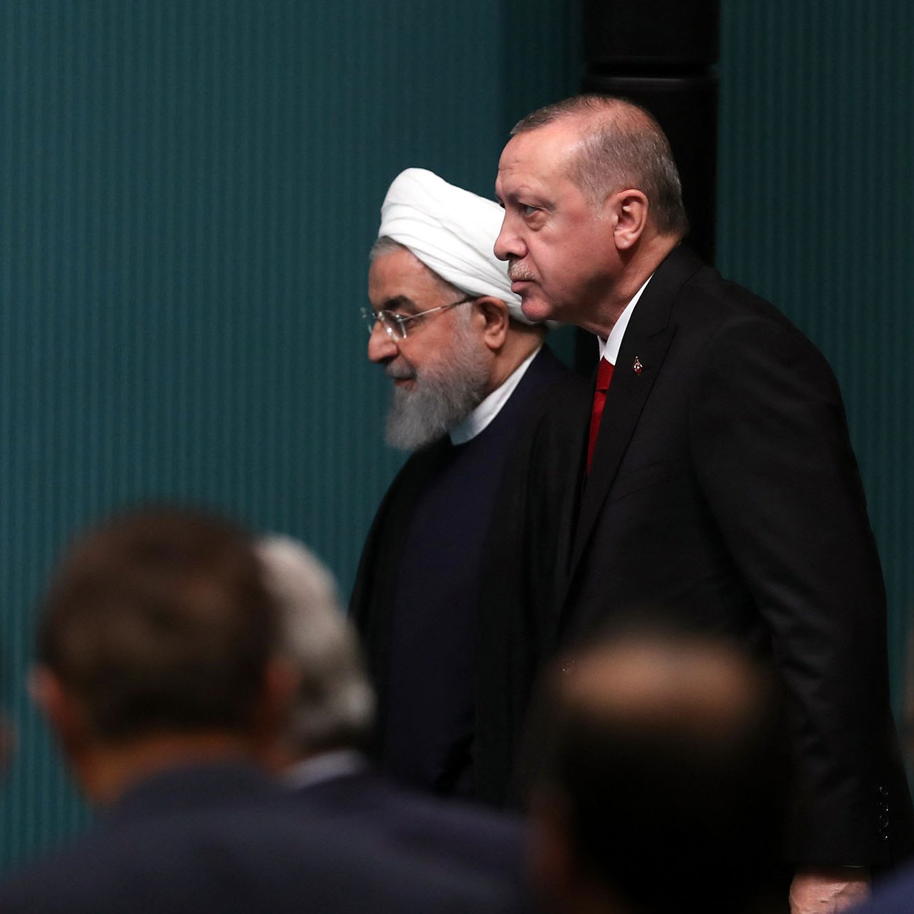 L'image montre deux hommes marchant côte à côte dans un cadre officiel. L'un porte un turban blanc et une robe traditionnelle, tandis que l'autre est habillé en costume avec une cravate. Ils semblent se diriger vers un événement, avec des personnes assises en arrière-plan. L'ambiance paraît formelle, suggérant une rencontre politique ou diplomatique.