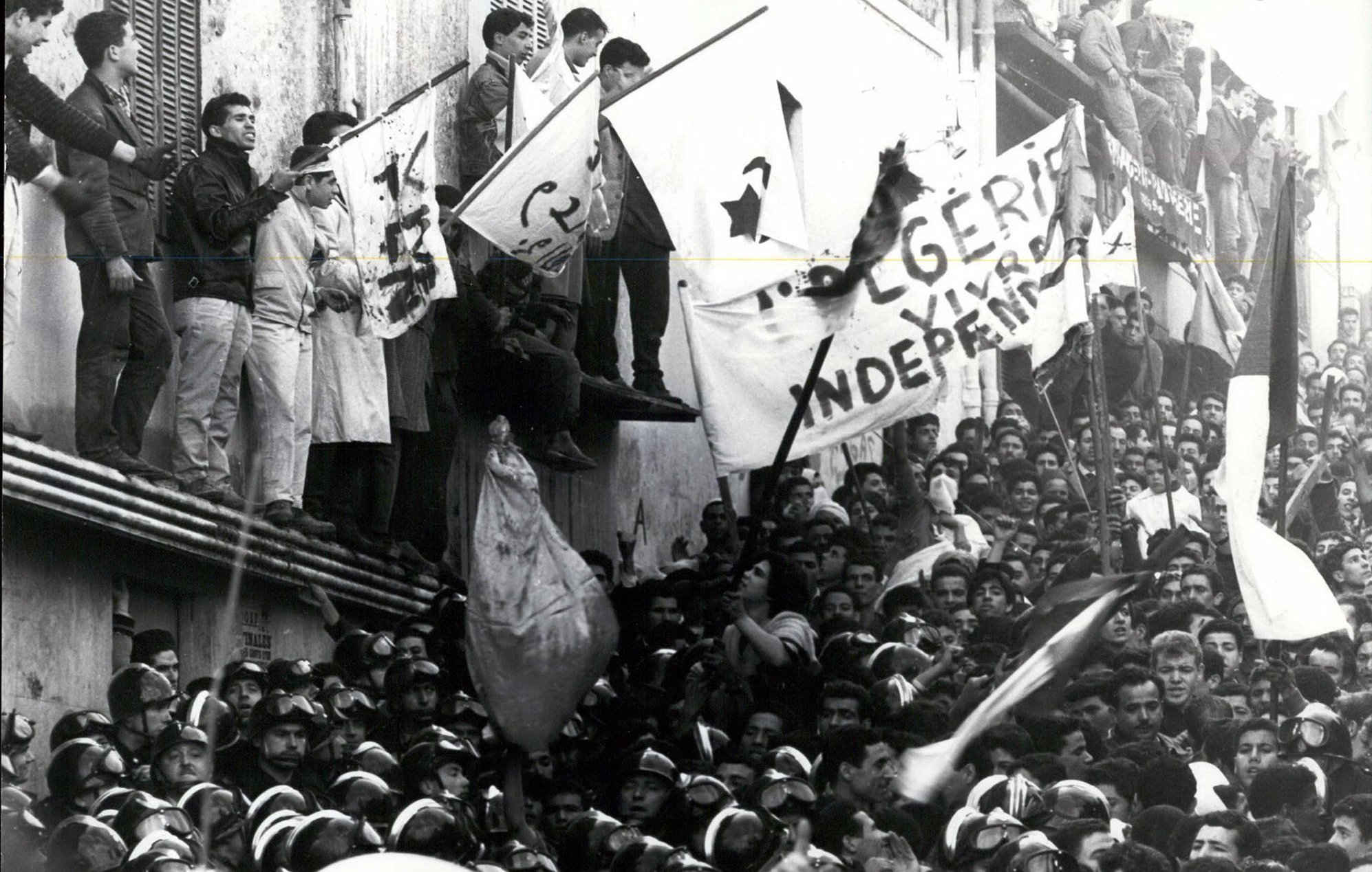 L'image montre une grande foule rassemblée, avec des individus brandissant des drapeaux et des bannières. L'atmosphère semble tendue et chargeée d'émotion, probablement liée à un événement historique. Des personnes se tiennent sur un bâtiment, tandis que d'autres sont regroupées en bas, certaines portant des uniformes, ce qui pourrait indiquer une présence policière ou militaire. Le message sur les bannières évoque des thèmes d'indépendance, suggérant un contexte de luttes sociales ou politiques.