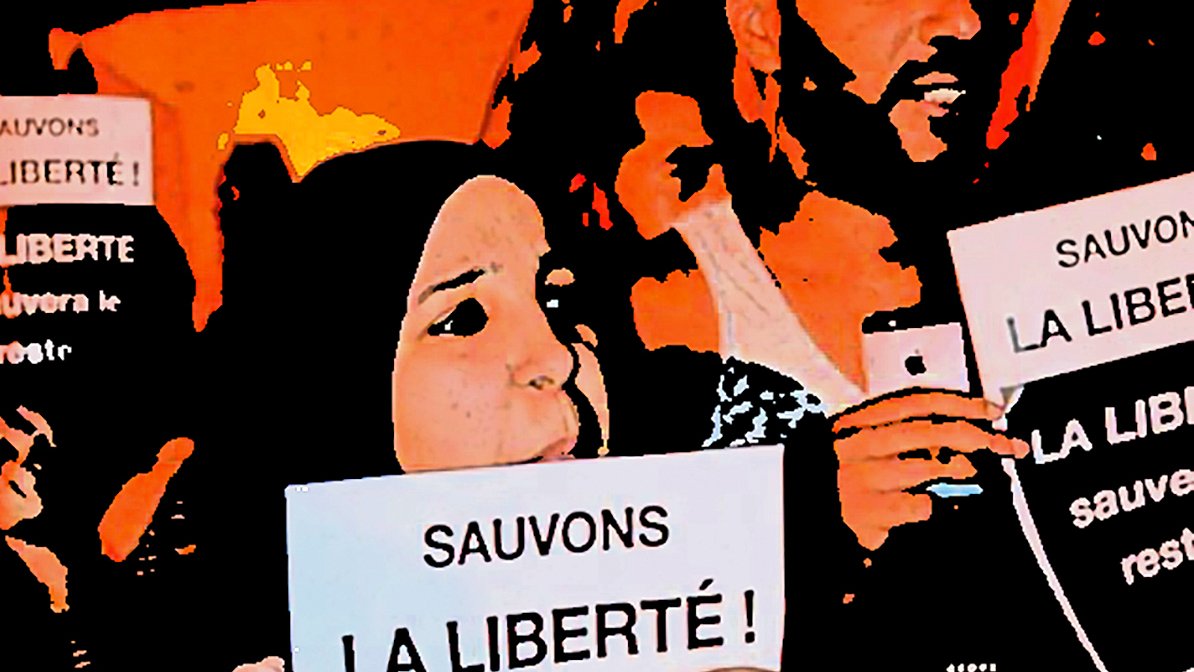 تظهر في الصورة مجموعة من الأشخاص المشاركين في احتجاج أو مظاهرة، حيث يحملون لافتات مكتوب عليها "لننقذ الحرية!" و"الحرية ستنقذ البقية...". يبدو من تعبيرات وجوههم أنهم متحمسون وملتزمون بالقضية التي يتظاهرون من أجلها. الخلفية تعكس أجواء حماسية ودعماً للقضية المطروحة.