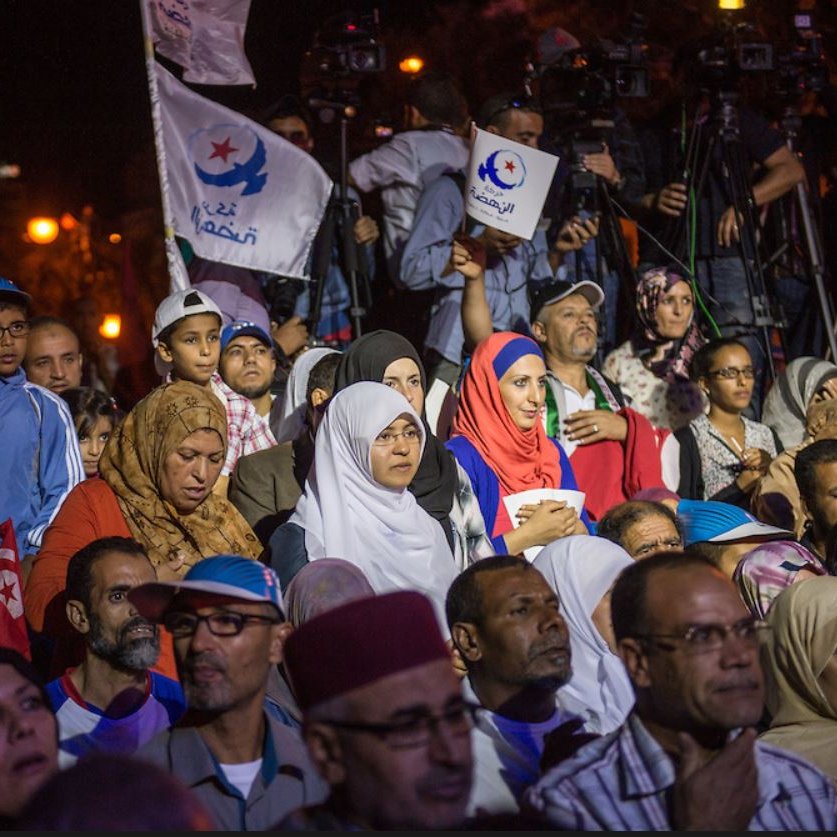L'image montre une foule rassemblée lors d'un événement, probablement politique. On peut voir des personnes de différents âges, y compris des enfants. Certaines personnes portent des drapeaux avec des symboles, suggérant qu'elles soutiennent un mouvement ou un parti. L'atmosphère semble intense, avec des caméras et des journalistes présents, indiquant l'importance de l'événement. Les gens affichent diverses expressions, allant de la concentration à l'enthousiasme.