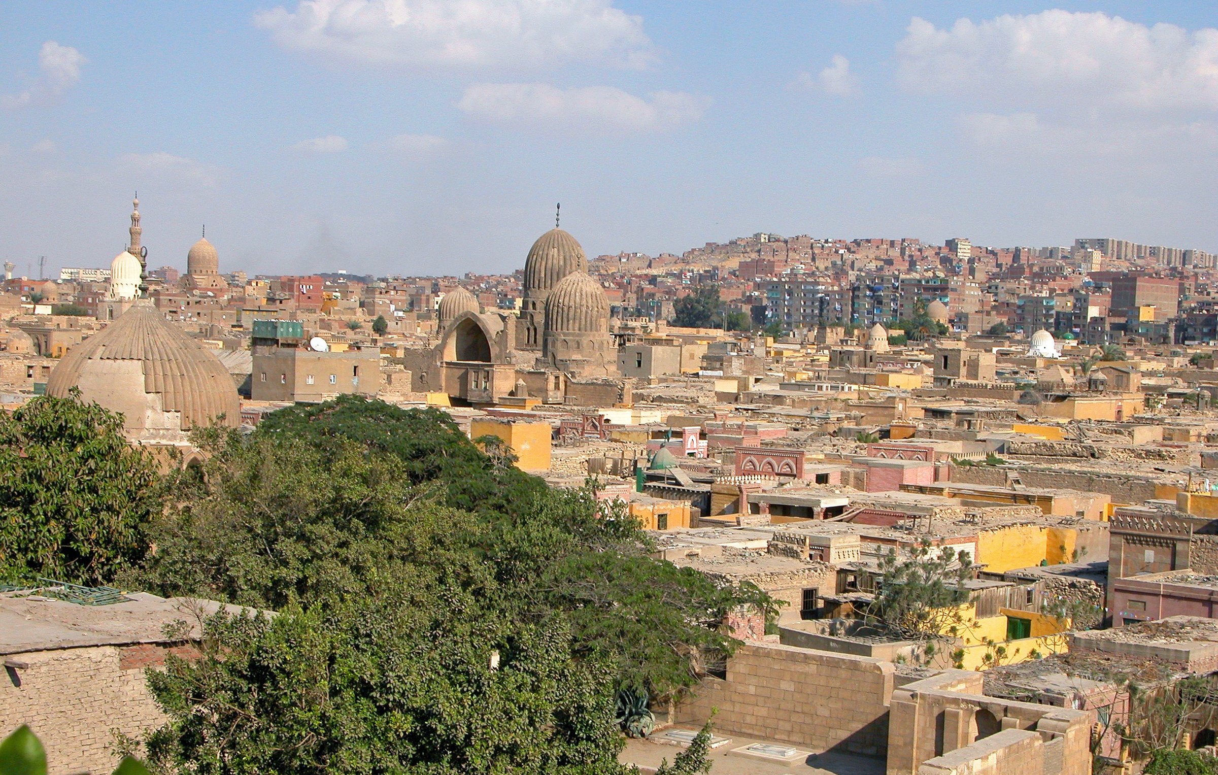 L'image montre une vue panoramique d'une ville historique, probablement un quartier ancien avec des bâtiments en pierre. On peut voir plusieurs coupoles et minarets qui témoignent de l'architecture traditionnelle. Les toits sont variés, certains sont plats et d'autres sont plus élevés, tandis que la végétation semble présente avec des arbres. En arrière-plan, on aperçoit des collines et d'autres bâtiments, suggérant une ville vaste et densément peuplée. Le ciel est clair avec quelques nuages, donnant une atmosphère lumineuse à la scène.