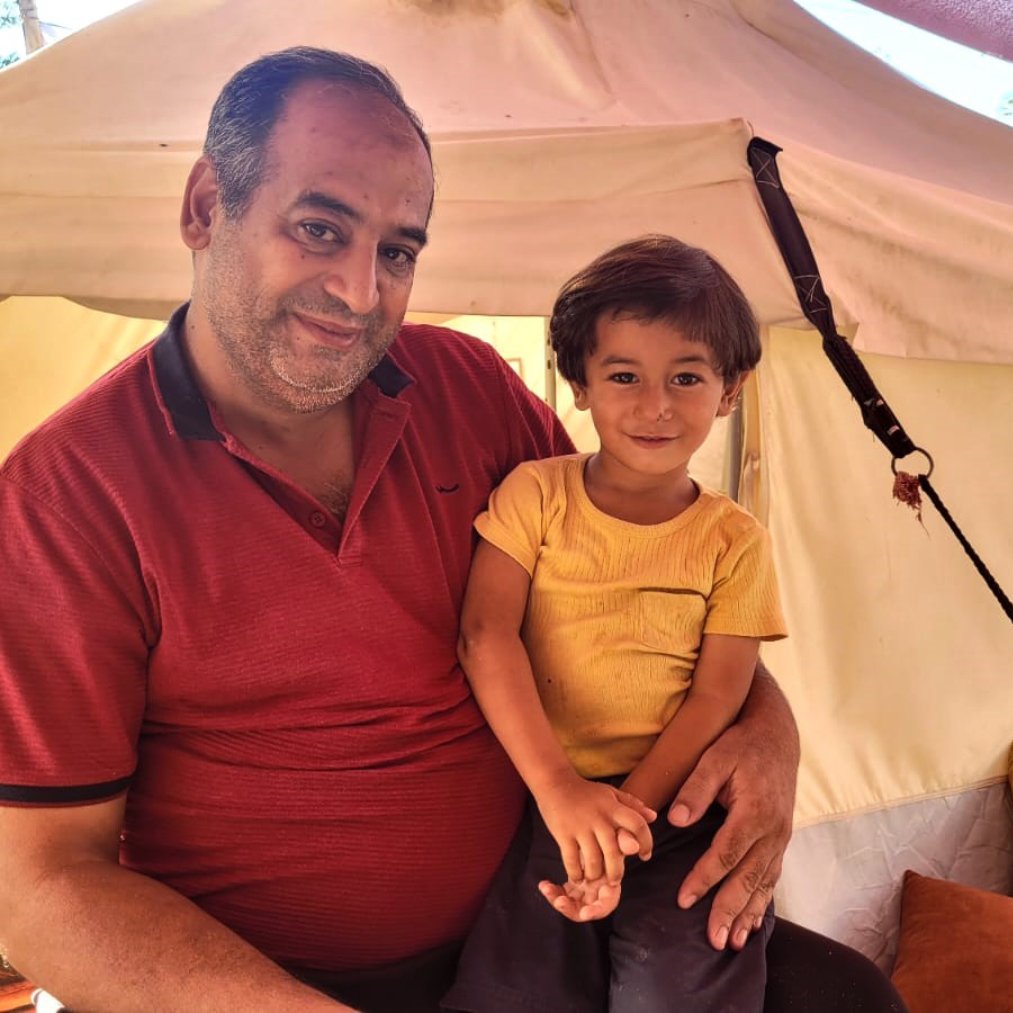 L'image montre un homme souriant assis à l'intérieur d'une tente, tenant un jeune enfant sur ses genoux. L'homme porte une chemise rouge et a l'air détendu et joyeux. L'enfant, qui porte un t-shirt jaune, sourit également et semble heureux. L'intérieur de la tente est simple, avec des coussins au sol, créant une atmosphère conviviale et chaleureuse.