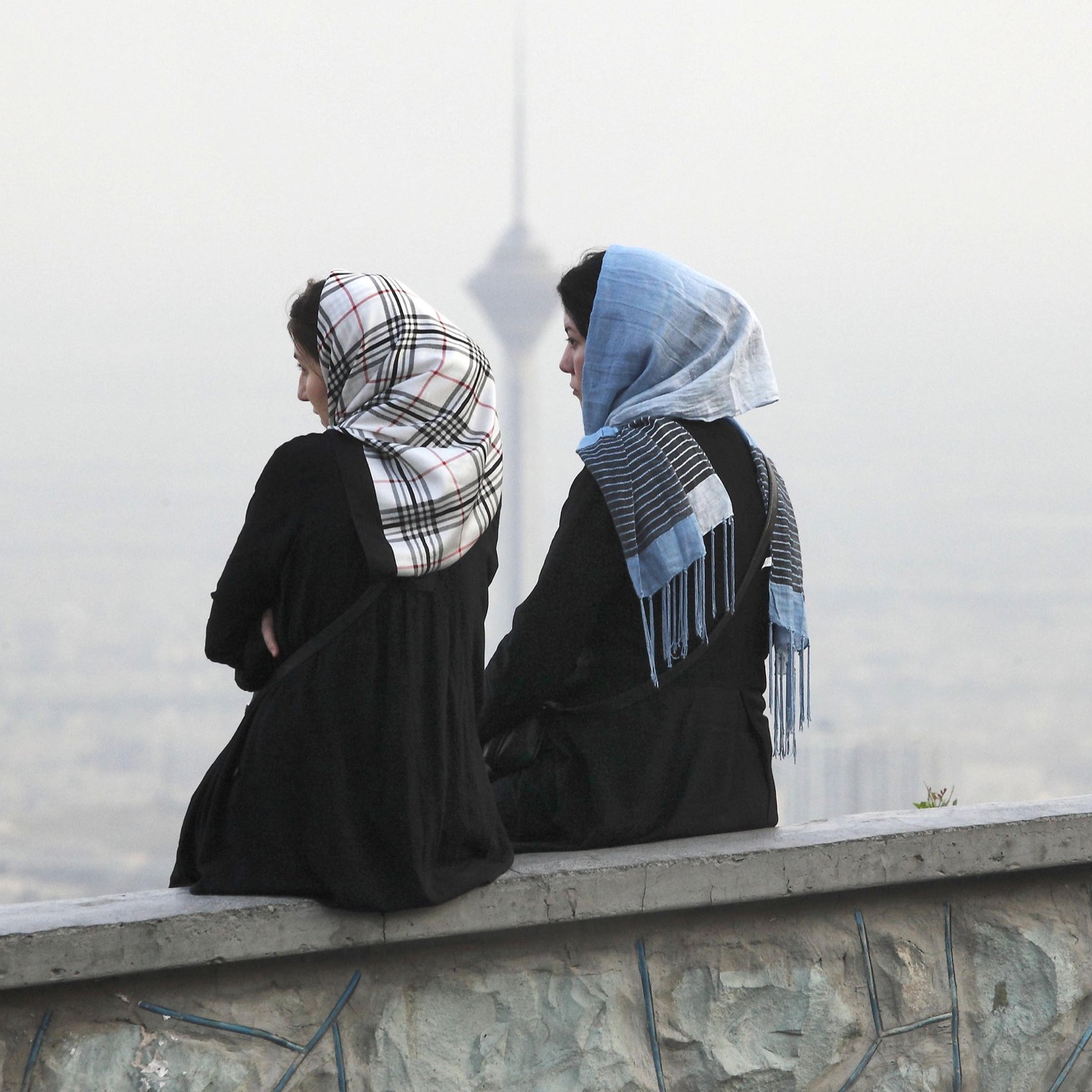 L'image montre deux femmes assises sur un mur, avec le dos tourné à la caméra. Elles portent des vêtements longs et des foulards, l'un d'eux étant à motifs. En arrière-plan, on aperçoit une ville embrumée avec ce qui semble être une tour, ajoutant un aspect urbain à la scène. L'atmosphère est paisible, avec une lumière douce, suggérant un moment de réflexion ou de contemplation.