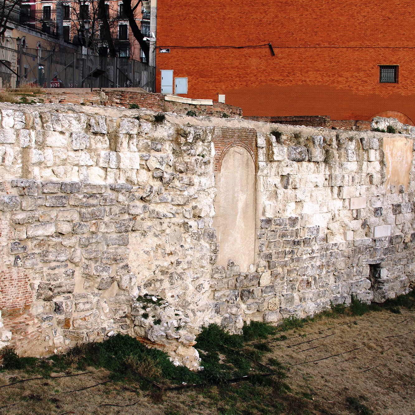 L'image montre une section de mur en pierre, probablement une ancienne structure ou fortification. Les pierres sont de tailles et de formes variées, avec des parties couvertes de végétation. En arrière-plan, on peut apercevoir un mur de couleur rouge et quelques éléments de structure moderne. Le site semble être à l'extérieur, probablement dans une zone historique, avec une ambiance de délabrement et d'abandon.