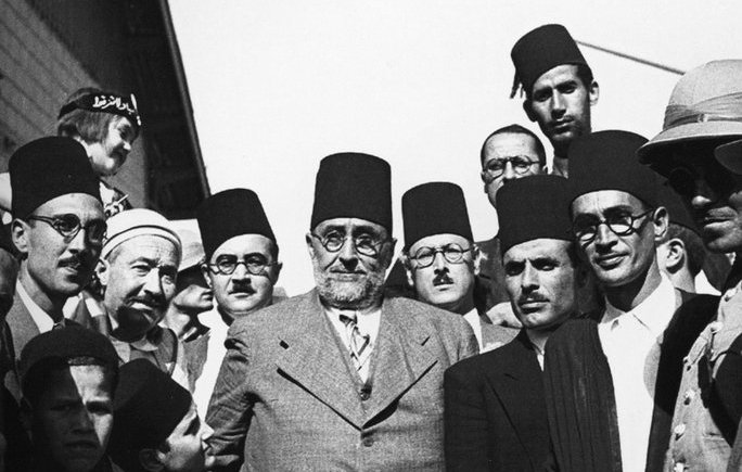 L'image représente un groupe d'hommes en costumes formels, majoritairement portant des fez. Ils semblent être engagés dans une discussion ou un moment de camaraderie. L'ambiance est sérieuse, et les expressions des visages montrent une concentration ou une détermination. Une femme est également visible en arrière-plan, portant un foulard. Le cadre pourrait indiquer un événement historique ou un rassemblement important, probablement dans un contexte culturel ou politique.