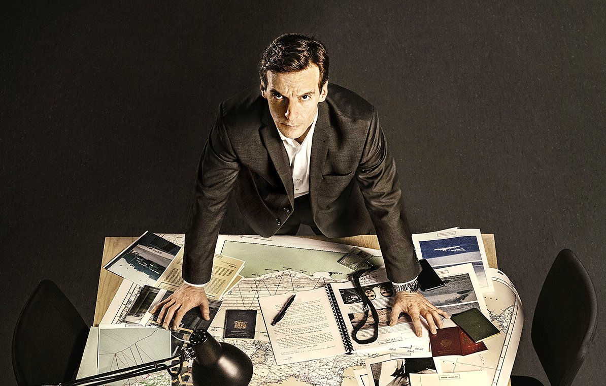 L'image montre un homme debout sur une table en bois, entouré de divers documents et d'objets. Il a une expression sérieuse et semble concentré. Sur la table, on peut voir des cartes, des notes, des dossiers et un lampadaire qui éclaire la scène. L'environnement autour de lui est plutôt sobre, avec des chaises noires et un sol sombre. L'ensemble donne une impression de travail acharné et de recherche.