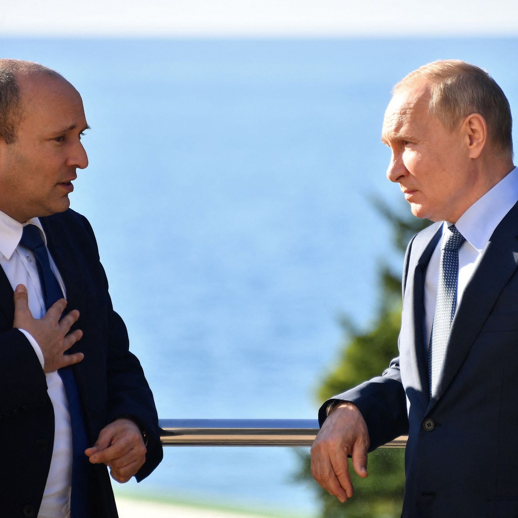 تظهر الصورة شخصين يتحدثان معًا، يبدو أن المحادثة جادة. أحدهما يرتدي بدلة رسمية مع ربطة عنق، بينما الآخر يرتدي أيضًا بدلة ولكن مع شارة دينية على رأسه. الخلفية تظهر منظرًا طبيعيًا جميلًا مع البحر. يُظهر كل منهما اهتمامًا بما يقوله الآخر، مما يوحي بأهمية النقاش بينهما.