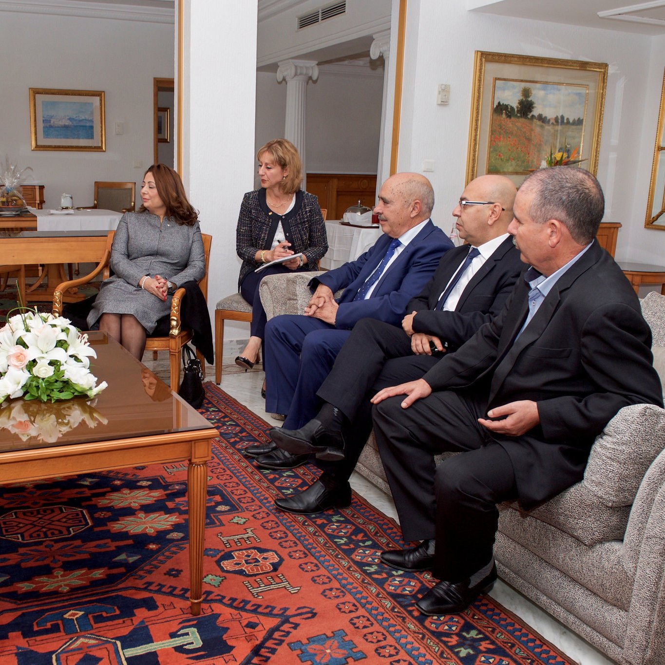 The image depicts a formal meeting taking place in a well-lit, elegantly furnished room. A group of professionals is seated around a coffee table, engaged in discussion. On one side, a person in a suit appears to be leading the conversation, while others listen attentively. The room features tasteful decor, including paintings on the walls and a floral arrangement on the table, creating a calm and sophisticated atmosphere. The group consists of both men and women, indicating a diverse representation.