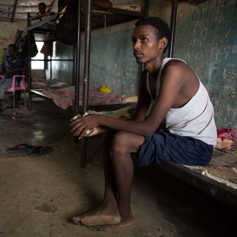 L'image montre un espace intérieur qui semble être une chambre ou un dortoir. On y voit plusieurs lits superposés et des personnes assises ou allongées. Un jeune homme est assis sur le bord d'un lit, regardant pensivement au loin. Il porte un débardeur et un short, et ses pieds sont nus. Les murs de la pièce sont nus et usés, avec des traces d'humidité, ce qui indique un environnement modeste et probablement surpeuplé. La lumière naturelle pénètre par une fenêtre, ajoutant une certaine ambiance à la scène.