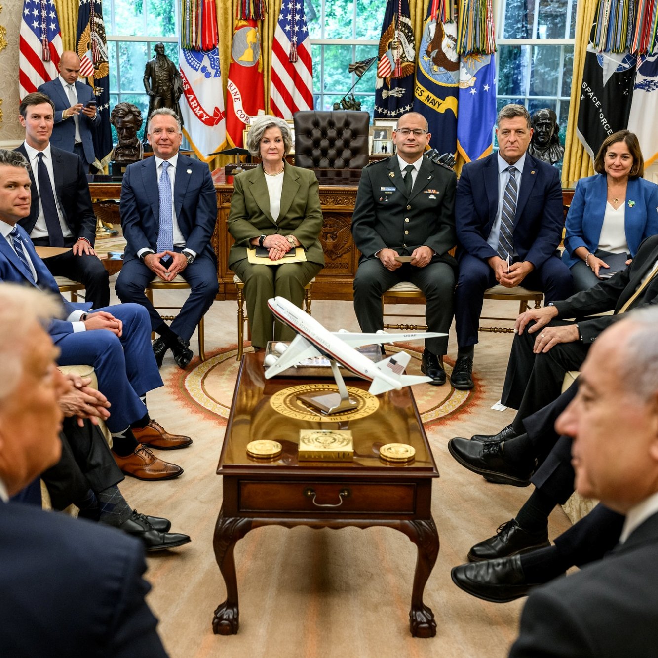In una sala con bandiere, vari uomini e donne seduti attorno a un tavolo con un aereo in miniatura.