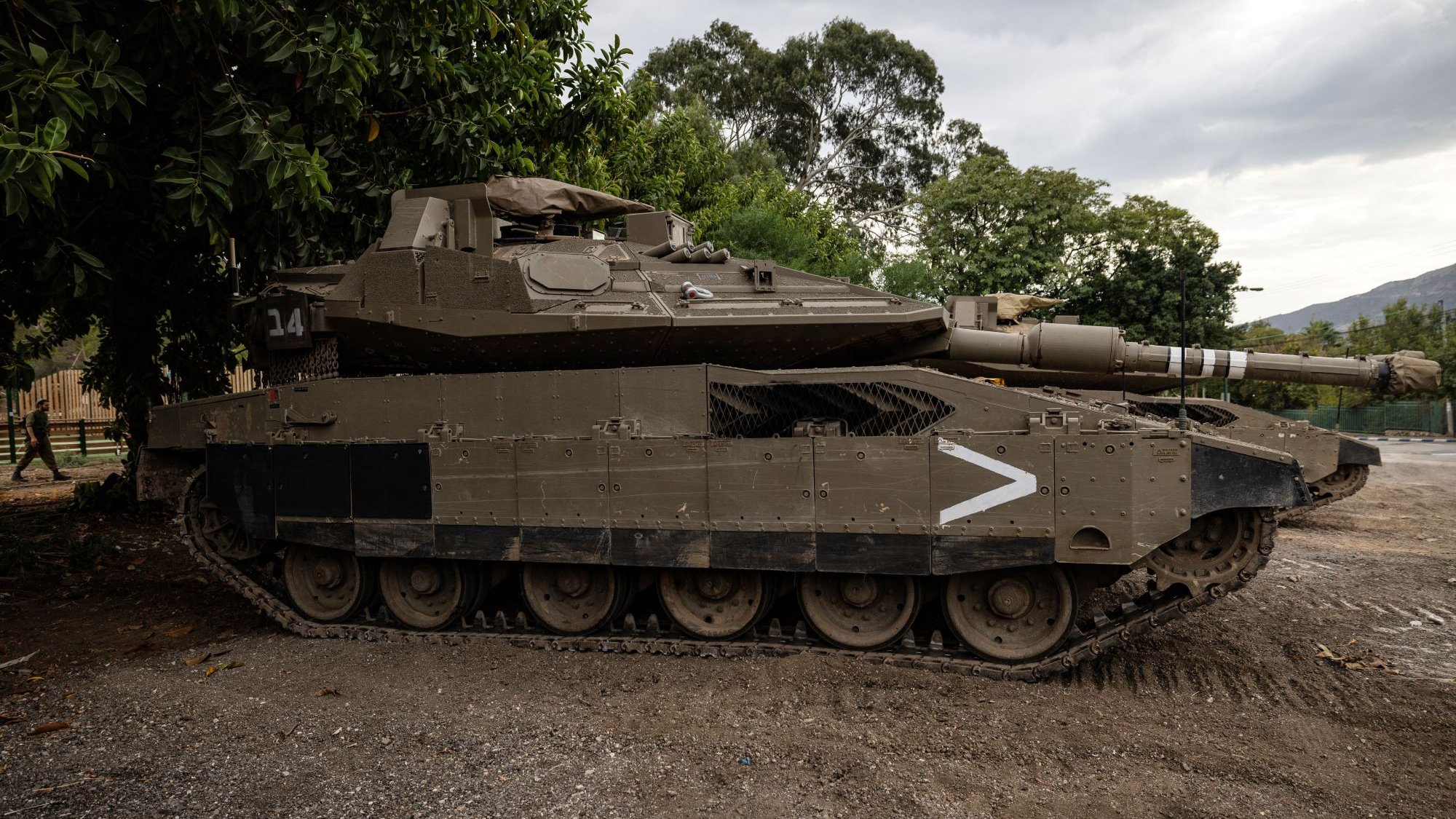 الصورة تظهر دبابة مدرعة على الأرض، متوقفة بجانب مجموعة من الأشجار. الدبابة تحمل لوناً زيتونياً وتبدو قوية ومتينة، مع تفاصيل واضحة مثل البرج والمدفع. السماء خلفها ملبدة بالغيوم، مما يعطي انطباعًا عن أجواء درامية. الإطار العام يوحي بمكان عسكري أو منطقة تدريب.