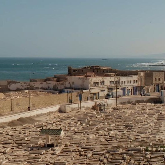 L'image montre un panorama d'une ville côtière, probablement située au bord de la mer Méditerranée. Au premier plan, on peut voir un cimetière avec de nombreuses tombes en pierre, disposées de manière régulière. À l'arrière-plan, il y a des bâtiments blancs, typiques des architectures méditerranéennes, qui bordent la côte. La mer s'étend à l'horizon, avec des vagues visibles, et il semble y avoir une légère brume sur l'eau. La scène dégage une ambiance paisible et sereine, avec une lumière douce évoquant l'heure du soir ou du matin.