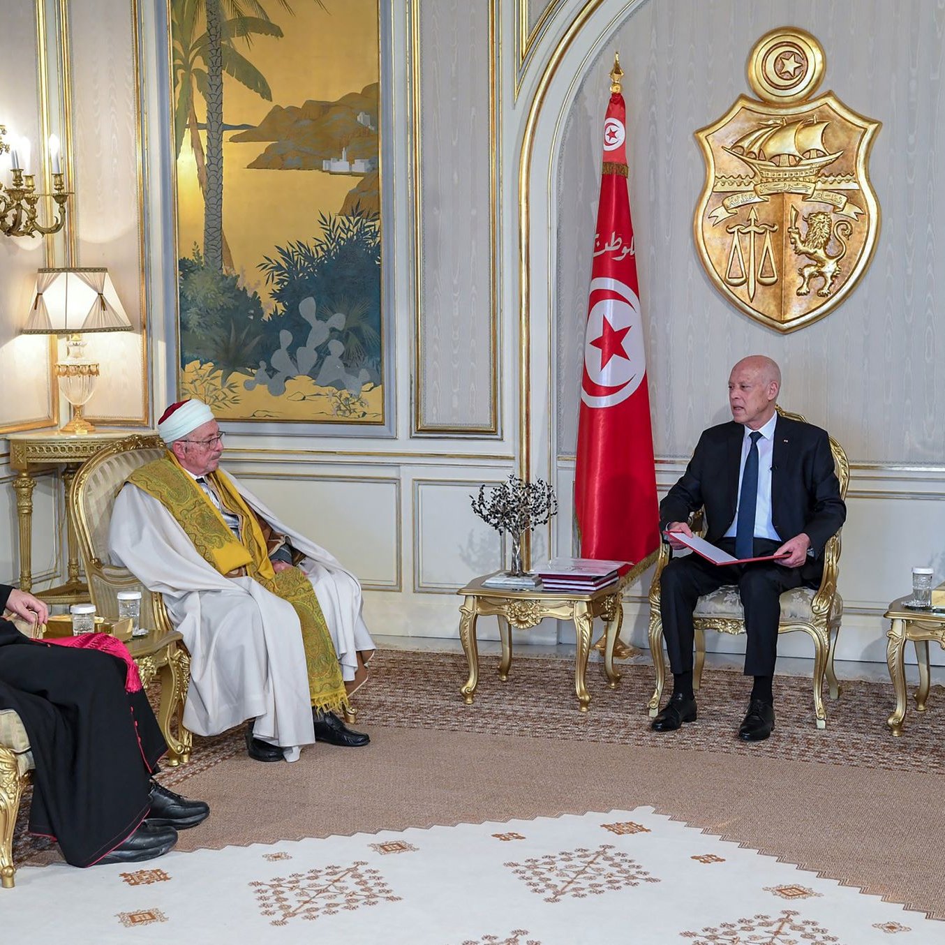 L'image montre une réunion dans un cadre formel. On peut voir plusieurs personnes assises autour d'une table basse. À gauche, un homme en soutane rouge, probablement un évêque, et un homme en robes traditionnelles, au centre, qui semble être un chef religieux. À droite, un homme en costume noir. Au fond, le drapeau tunisien flotte, et on peut apercevoir des éléments de décoration élégants sur les murs et des plantes en arrière-plan. La scène évoque un moment de dialogue interreligieux ou de rencontre officielle.