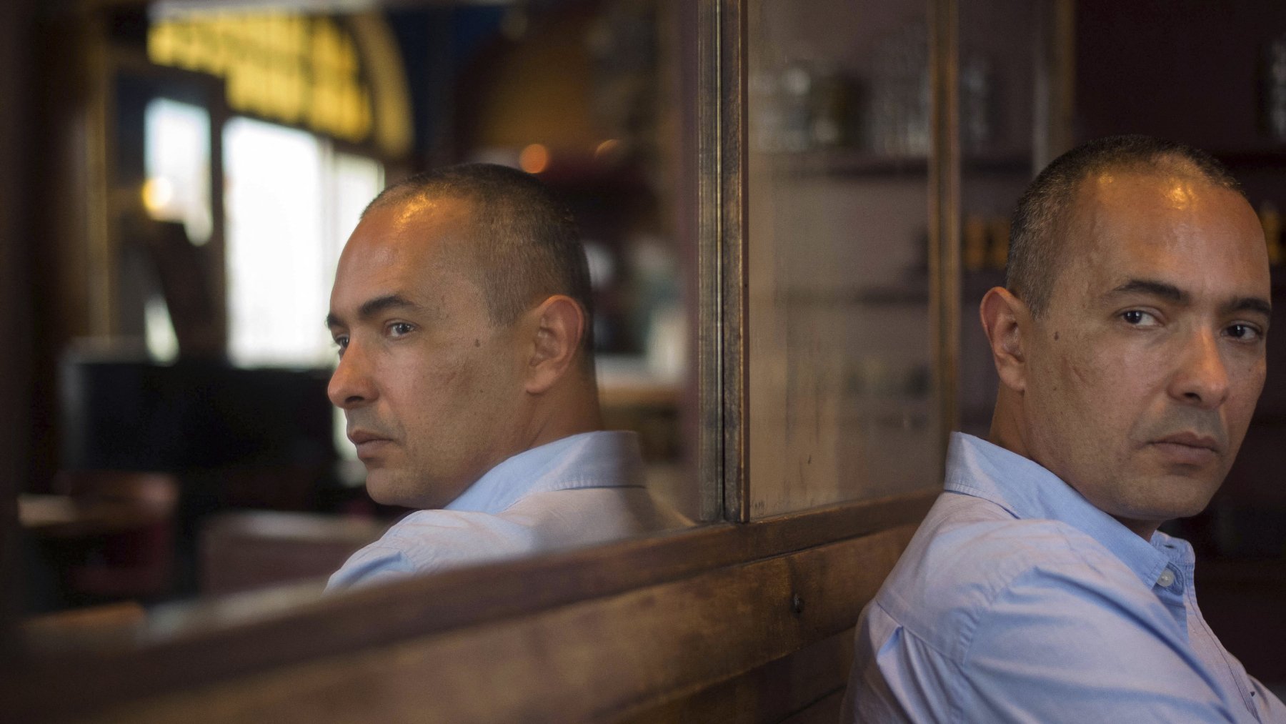 تظهر الصورة شخصًا يجلس على مقعد خشبي، مع انعكاسه في زجاج خلفه. يبدو أن الرجل في منتصف العمر، لديه شعر قصير ومظهر هادئ. يرتدي قميصًا أزرق فاتح، وينظر مباشرة إلى الكاميرا بتعبير جاد. الخلفية غير واضحة، مما يركز الانتباه على الشخص ووجهه. الإضاءة في الصورة دافئة وتساهم في خلق جو من التأمل والعمق.