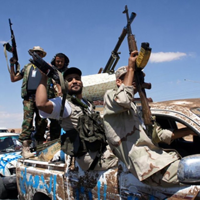 L'image montre des hommes armés debout sur un véhicule, probablement un pick-up, dans un environnement désertique ou semi-désertique. Ils affichent des signes de victoire ou de célébration, brandissant leurs armes au-dessus de leur tête. Le véhicule est couvert de boue et décoré de drapeaux ou d'insignes. Le ciel est dégagé, suggérant une journée ensoleillée. L'ensemble donne une impression de mouvement et de dynamisme, avec un contexte de conflit ou de célébration militaire.