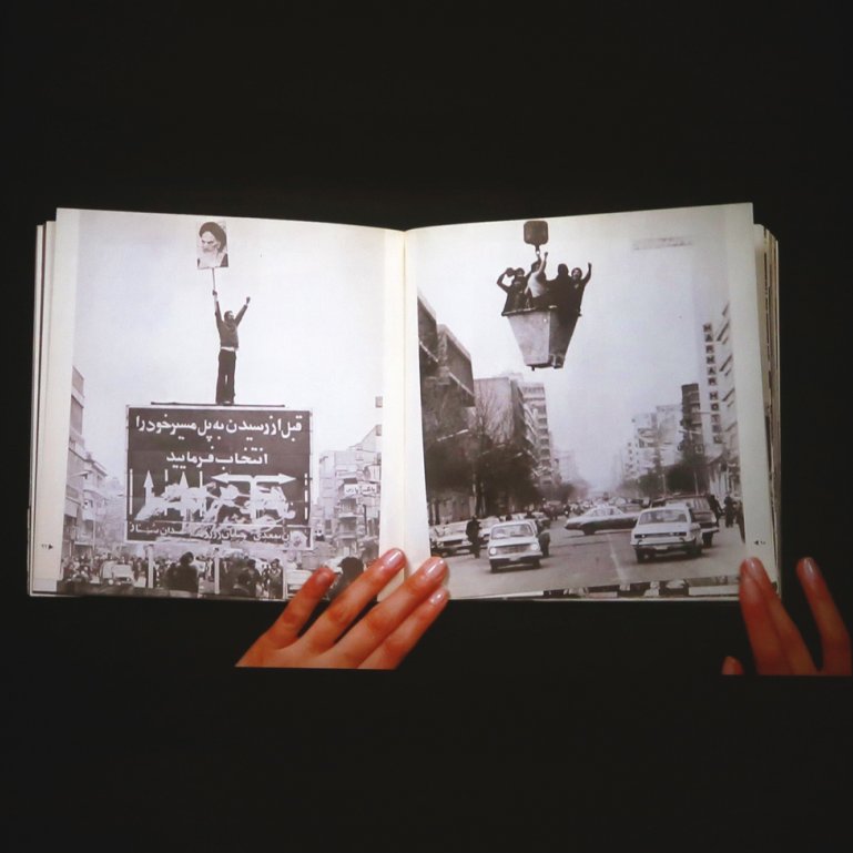 L'image montre un livre ouvert, tenu par des mains, avec des pages affichant des photos en noir et blanc. L'une des pages présente une grande pancarte ou affiche, tandis que l'autre illustre une rue avec des voitures. L'ensemble évoque des thèmes de mouvement, de protestation ou d'événements historiques, en jouant sur le contraste entre les images et le texte visibles. L'arrière-plan est sombre, ce qui met en valeur le livre et les illustrations qu'il contient.