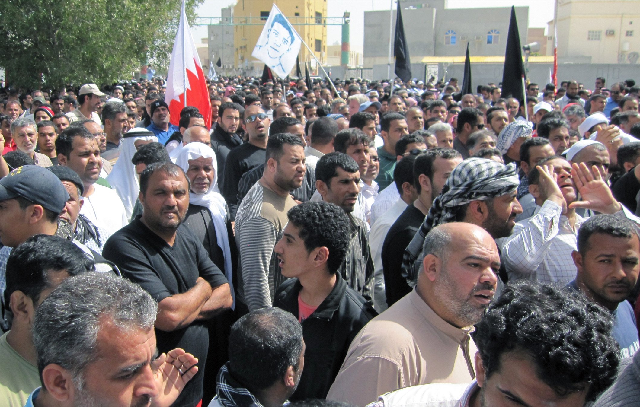 L'image montre une grande foule de personnes rassemblées, probablement lors d'une manifestation. Les participants semblent très engagés, certains tenant des drapeaux, y compris celui de Bahreïn. Les visages expriment diverses émotions, allant de la détermination à l'interrogation. L'environnement montre des bâtiments en arrière-plan, suggérant une zone urbaine. L'atmosphère générale dénote une forte implication politique ou sociale.