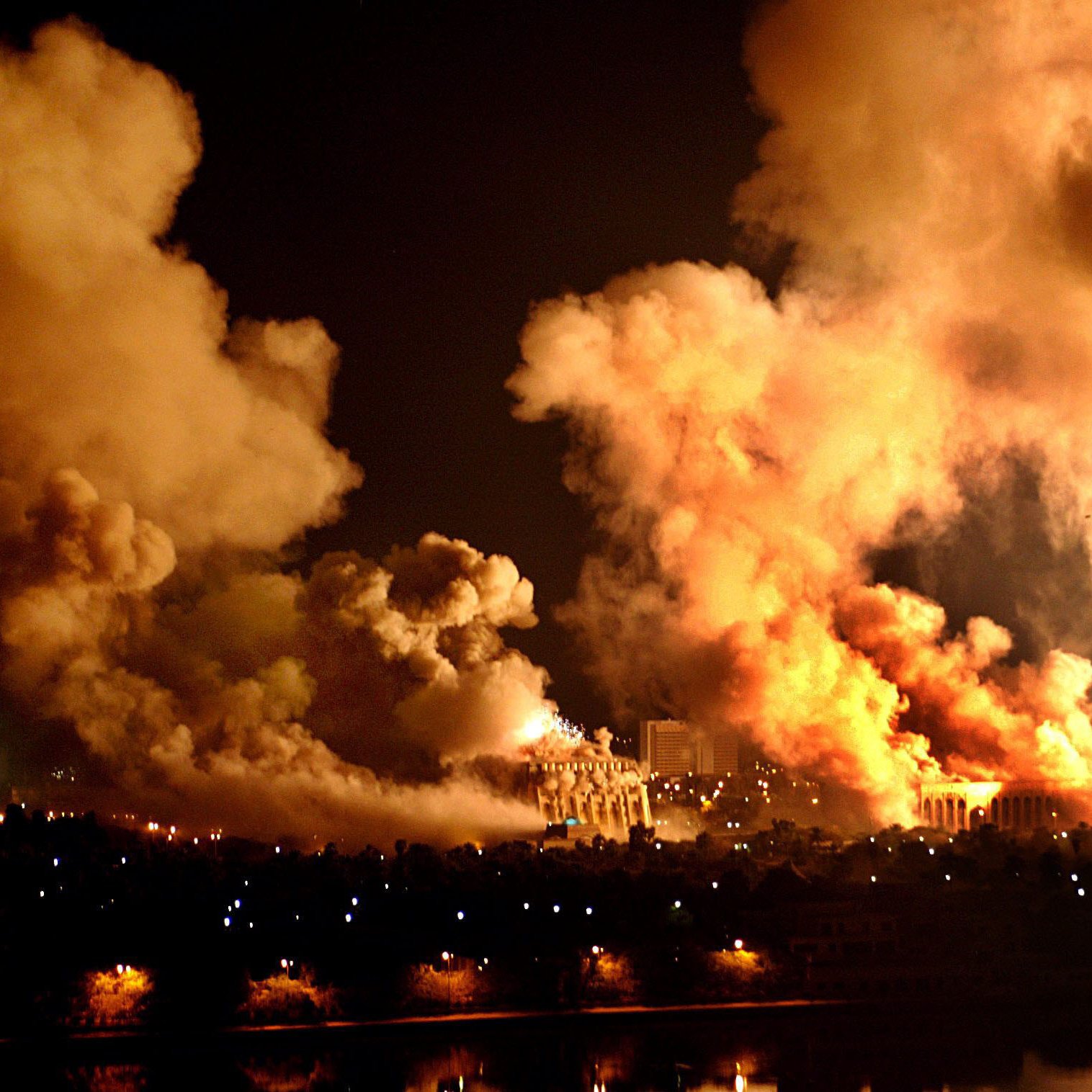 L'image montre une scène nocturne marquée par d'épais nuages de fumée et des flammes. On peut apercevoir des explosions ou des incendies créant une lumière intense dans l'obscurité. Les perturbations dans le ciel semblent témoigner d'un événement violent, tandis qu'en bas, on distingue une ville avec des lumières qui contrastent avec le chaos. L'ensemble dégage une atmosphère dramatique et inquiétante.