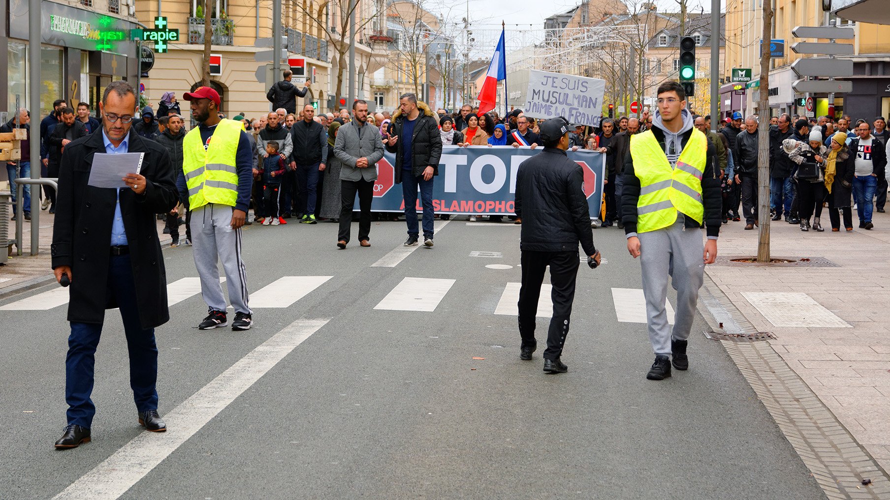 الصورة تظهر مجموعة من الناس يتظاهرون في شارع. في المقدمة، هناك شخصان يرتديان سترة عاكسة، ويبدو أنهما يجريان أو يقومان بمراقبة المظاهرة. في الخلفية، يمكن رؤية لافتة كبيرة مكتوب عليها "STOP ISLAMOPHOBIE"، مما يشير إلى أن المظاهرة تتعلق بمناهضة الإسلاموفوبيا. المتظاهرون يحملون لافتات ويبدو أنهم يتجمعون بشكل منظم. الجو العام يعكس النشاط والحيوية.