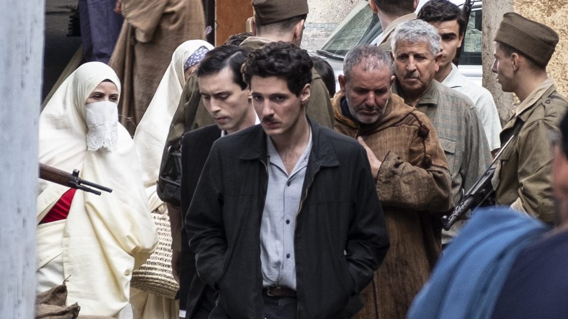 L'image montre une scène urbaine où plusieurs personnes sont rassemblées. Au centre, on voit un jeune homme en chemise claire et veste sombre, l'air pensif. Il est entouré de groupes de personnes, dont certaines semblent discuter ou se déplacer. Il y a également des hommes en uniformes militaires et des femmes portant des vêtements traditionnels, y compris un couvre-chef. L'ambiance de la scène semble chargée d'émotion, et le décor évoque une atmosphère historique ou dramatique.