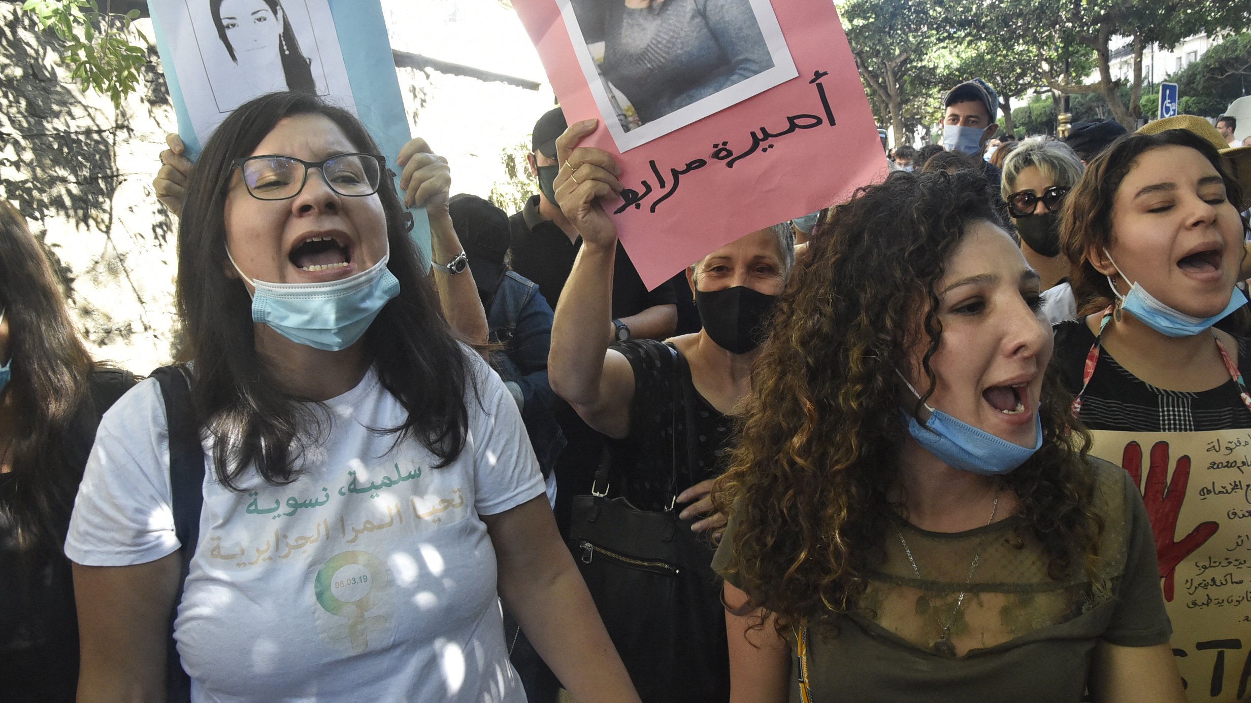L'image montre un groupe de manifestants lors d'une manifestation. Plusieurs personnes tiennent des pancartes avec des photographies et des slogans. Les participants semblent exprimés des émotions fortes, comme la colère ou la détermination, en scandant des slogans. Certaines personnes portent des masques de protection, tandis que l'environnement semble urbain avec des arbres et des bâtiments en arrière-plan.