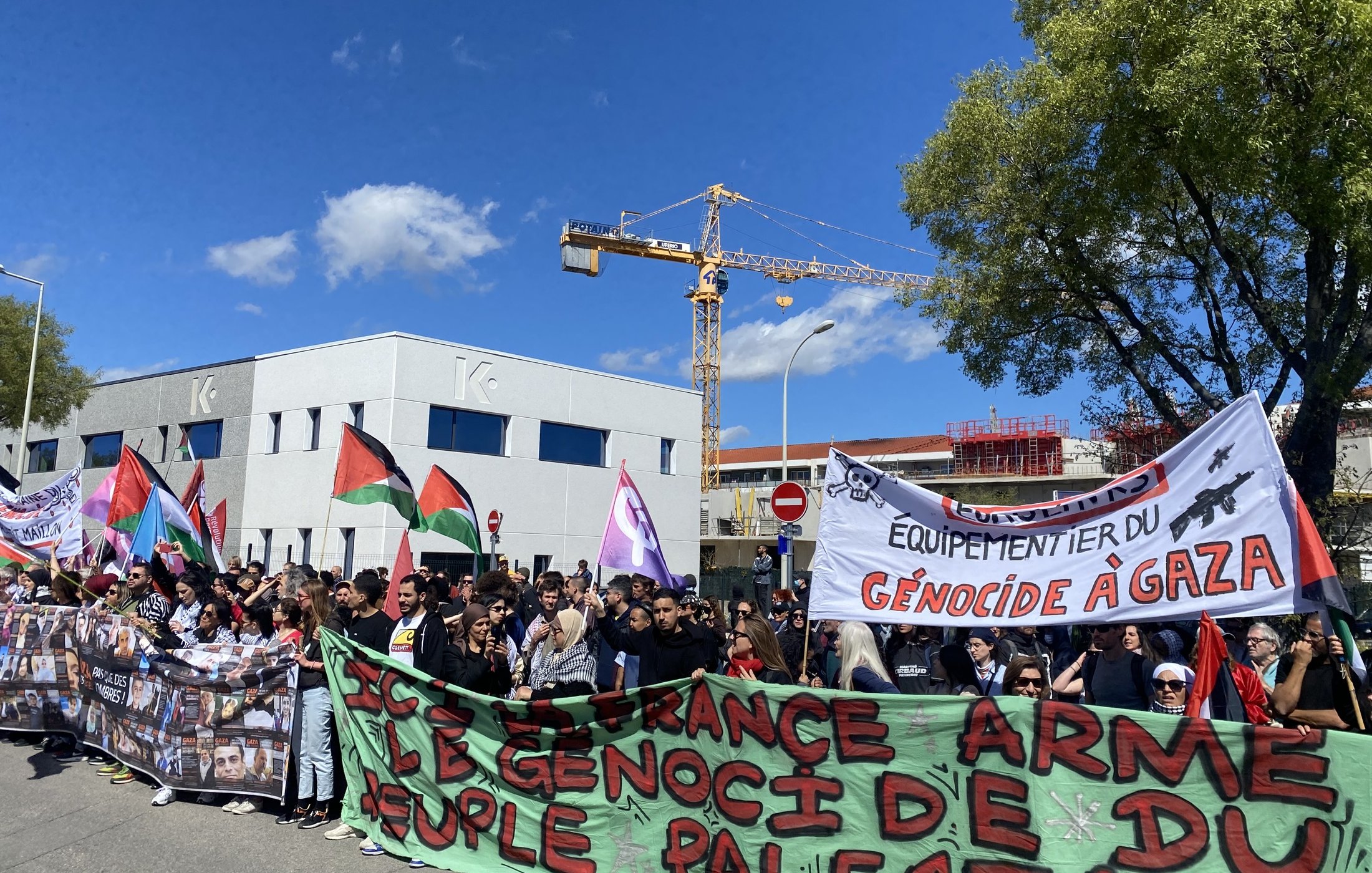 L'image montre une manifestation avec une grande foule de personnes rassemblées. Les manifestants portent des bannières et des drapeaux, y compris des drapeaux palestiniens. Certains panneaux affichent des messages en faveur des droits du peuple palestinien et dénoncent le génocide à Gaza. L'ambiance semble être celle d'une protestation pacifique, avec des participants unis pour une cause commune. En arrière-plan, on peut voir des bâtiments et une grue de construction.