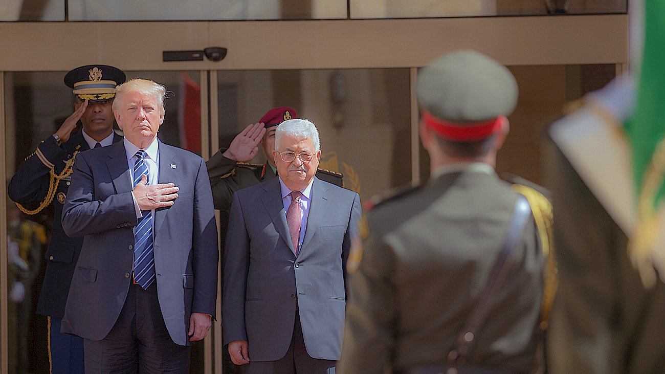 L'image montre deux hommes en tenue formelle, l'un à gauche avec une main sur le cœur et l'autre à droite debout, en présence d'une escorte militaire. Ils semblent participer à une cérémonie officielle. À l'arrière-plan, on peut voir un groupe de soldats en uniforme qui saluent. L'environnement suggère un cadre diplomatique ou d'État.