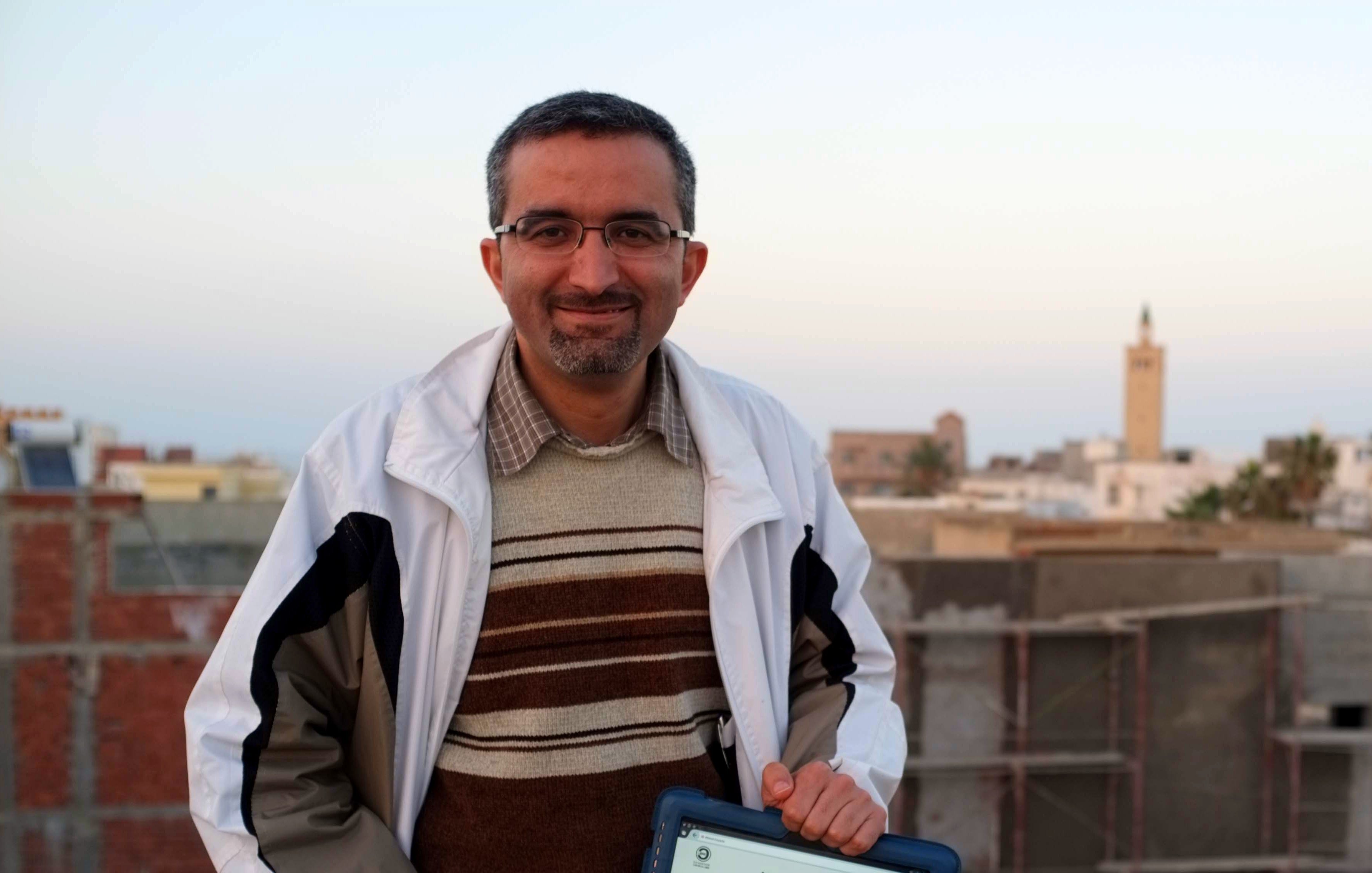L'image montre un homme souriant debout sur un toit, tenant une tablette. Il porte une veste blanche et un pull rayé. En arrière-plan, on peut voir des bâtiments et un minaret au loin, indiquant un paysage urbain. L'éclairage suggère un moment de la fin de journée, avec un ciel aux teintes douces.