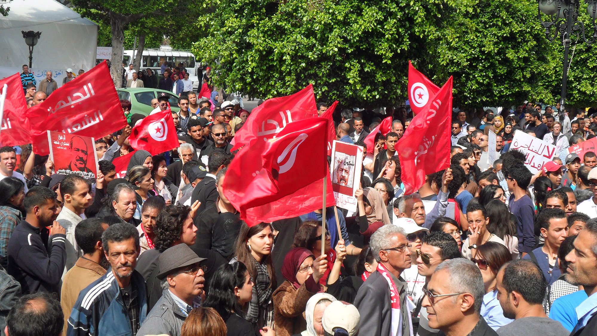 L'image montre une foule rassemblée dans une rue, lors d'une manifestation. Les participants tiennent des drapeaux rouges, probablement portant des symboles ou des slogans liés à la Tunisie. Il y a une grande variété de personnes dans la foule, de différents âges et expressions. L'ambiance semble énergique, avec des signes d'engagement et de participation active. En arrière-plan, on peut voir des bâtiments et des arbres, ce qui indique que l'événement se déroule dans un endroit urbain.