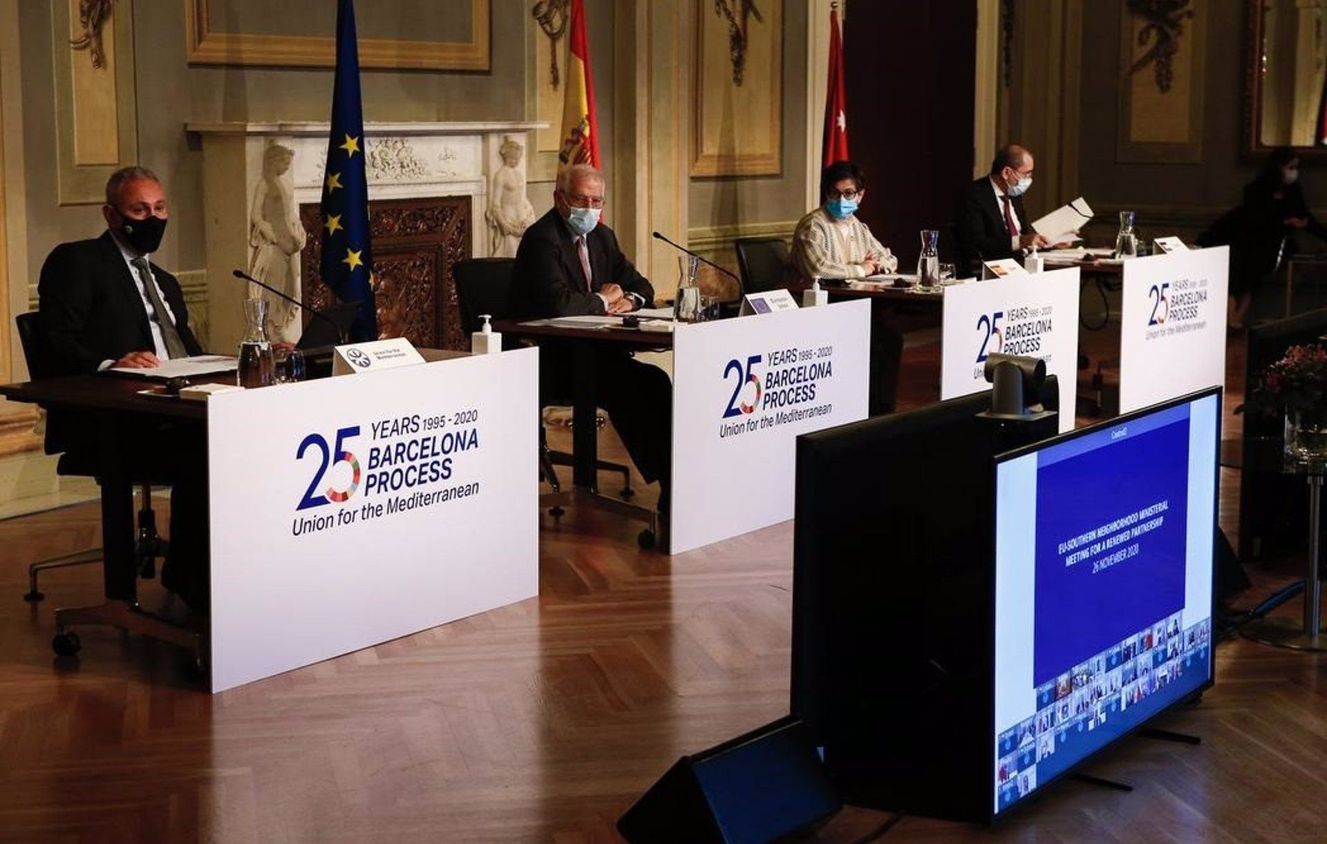 L'image montre une scène de conférence avec plusieurs intervenants assis derrière des panneaux portant le logo du processus de Barcelone, qui célèbre ses 25 ans. Les participants portent des masques, indiquant qu'ils suivent des mesures de santé, probablement en raison de la pandémie. À l'arrière-plan, on peut voir des drapeaux représentant l'Union européenne et l'Espagne, ainsi qu'un écran affichant des images, probablement liées à l'événement. L'environnement semble formel, suggérant une réunion diplomatique ou un sommet.