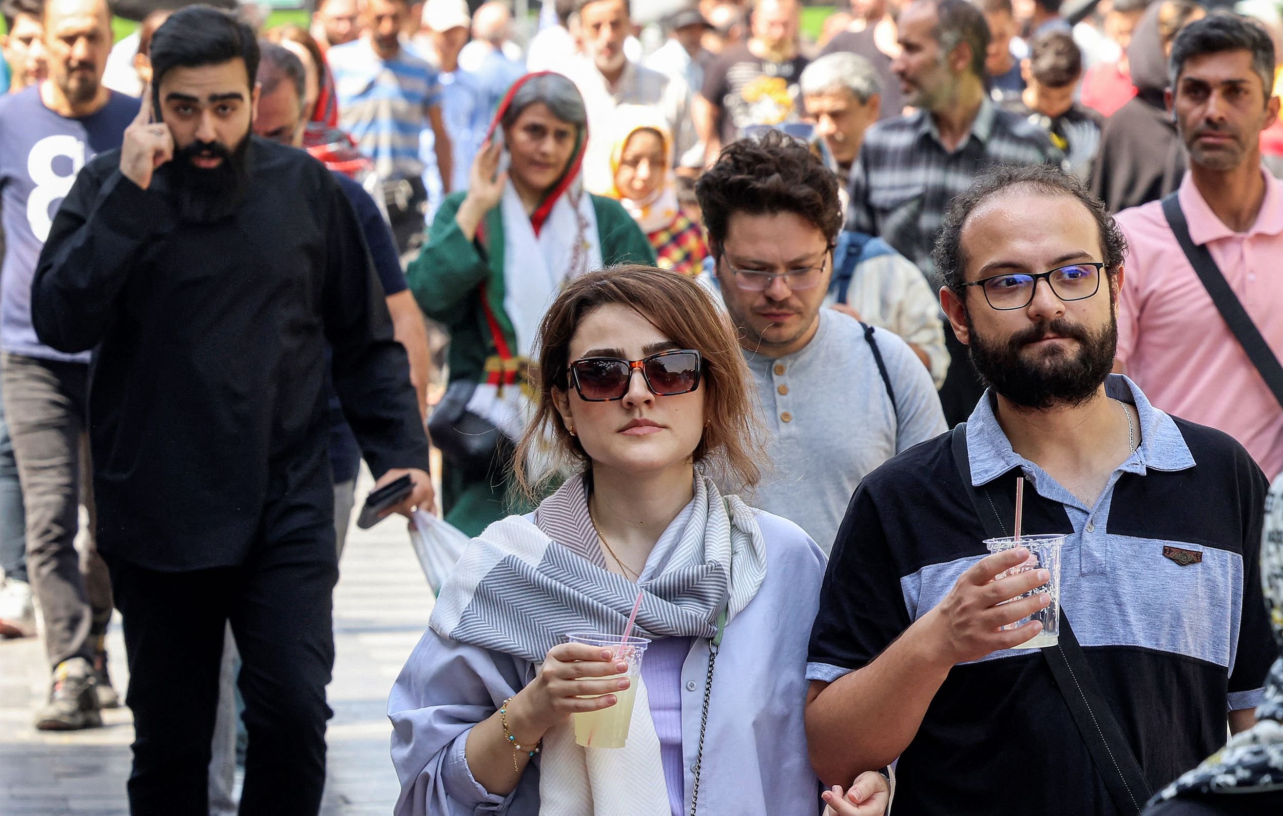 الصورة تظهر مجموعة من الأشخاص يسيرون في الشارع. في المقدمة، هناك امرأتان تحملان مشروبًا، بينما خلفهن يوجد رجال ونساء آخرون في مجموعات صغيرة. يبدو أن هناك تنوعًا في الأزياء بين الأشخاص، مع بعضهم يرتدي الملابس التقليدية والبعض الآخر يرتدي ملابس حديثة. الجو يبدو مفعمًا بالحركة والنشاط، مما يعكس أجواء الحياة اليومية في المدينة.