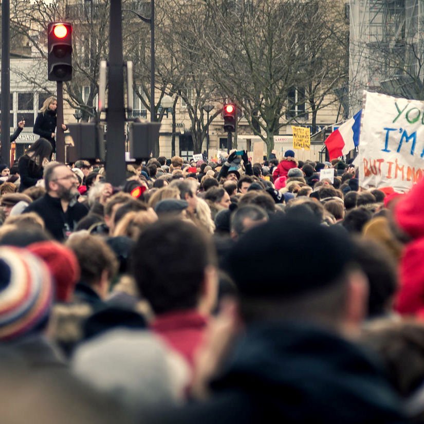 L'image montre une grande foule rassemblée, probablement pour une manifestation ou une marche. On peut voir des personnes de tous âges, certaines portant des écharpes ou des bonnets, tandis que d'autres sont vêtues de manteaux. Au centre, il y a une pancarte sur laquelle est écrit : "YOU MAY SAY I'M A DREAMER BUT I'M NOT THE ONLY ONE". Des drapeaux français sont visibles, ce qui suggère un contexte patriotique ou une revendication sociale. L'atmosphère semble être à la fois solide et engagée.